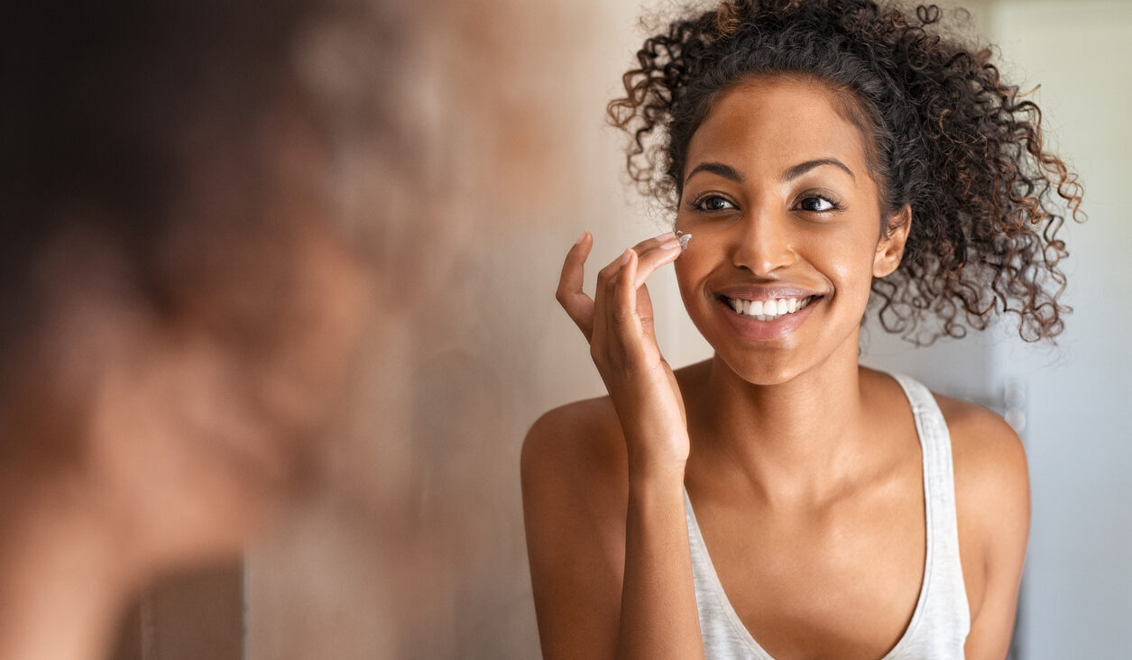 Young woman applying moisturising cream to her skin while standing in front of the mirror in the bathroom. Girl applying face cream while smiling. Beauty hydrating moisturizer and skincare routine concept.